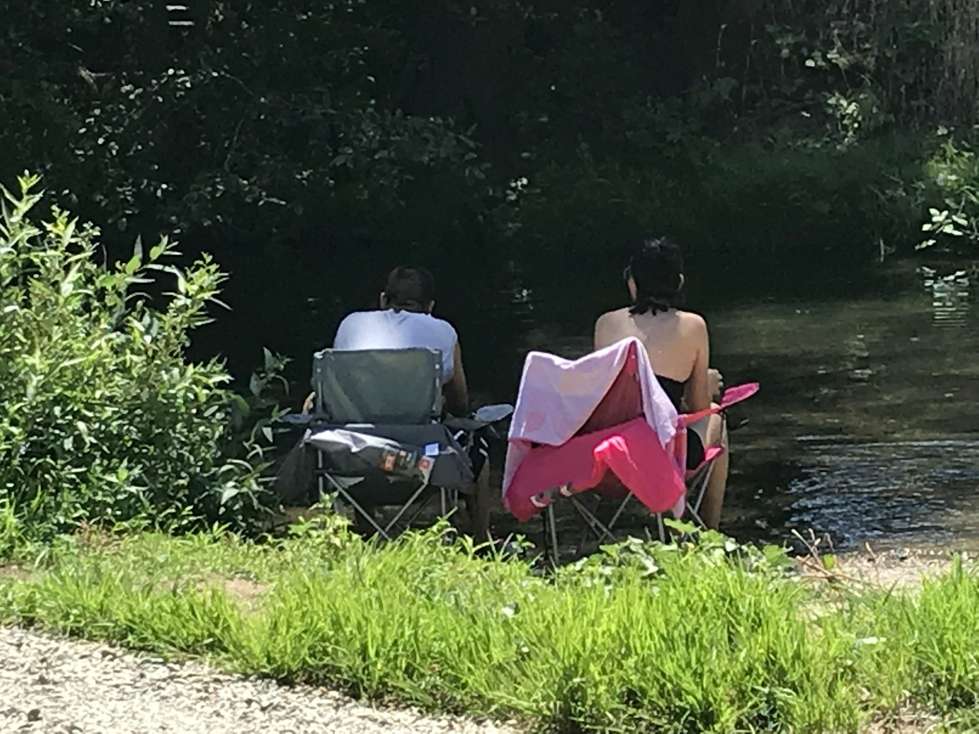Two people sitting in chairs by a riverside, surrounded by greenery.