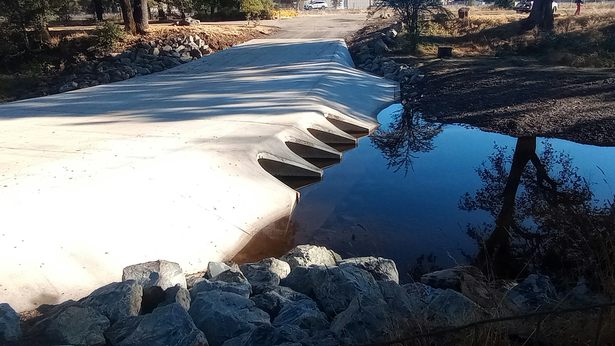 Concrete bridge over a small body of water with stone embankments, surrounded by trees and a parked car in the distance.
