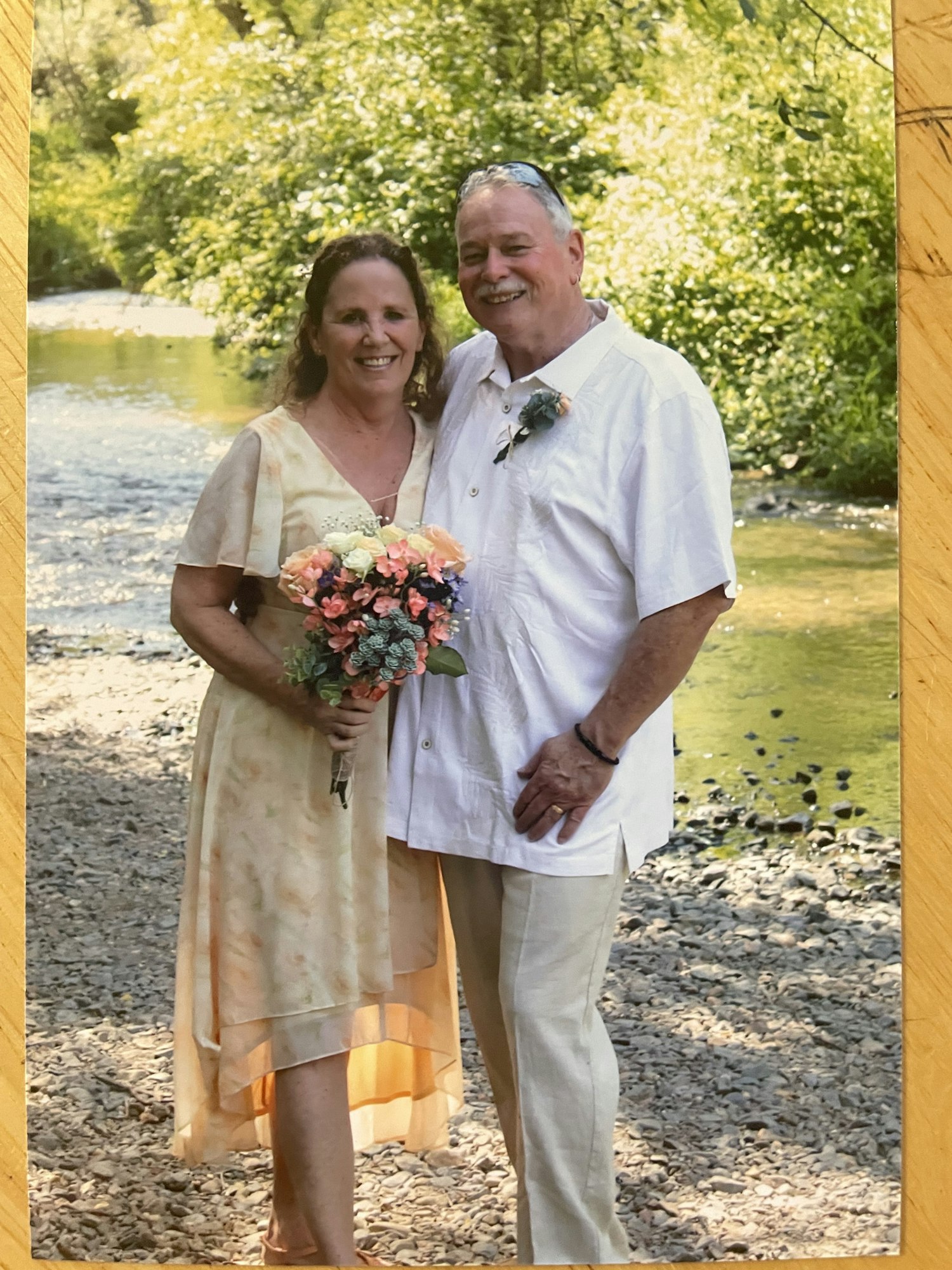A newly married couple smiling, standing by Squirrel Creek, with greenery in the background. The woman holds a bouquet, both are dressed casually.