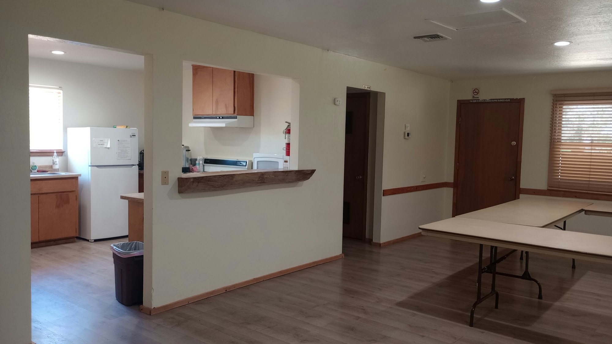 A room with a kitchen area, tables, refrigerator, cabinets, microwave, fire extinguisher, and a window with blinds.