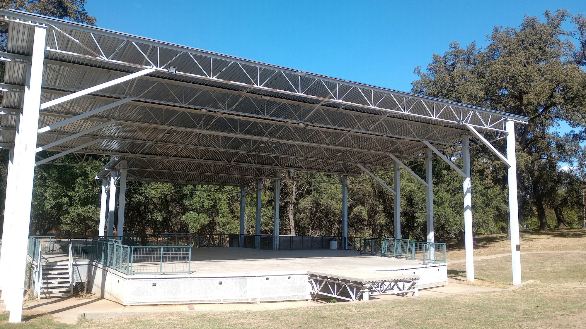 Outdoor stage with a metal roof, surrounded by trees, and a lawn in front.