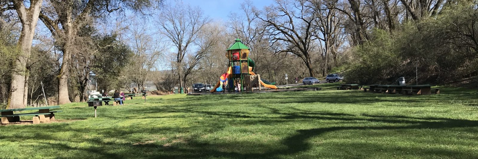 Playground with colorful slides, trees surrounding, benches, people, and parked cars on a sunny day.