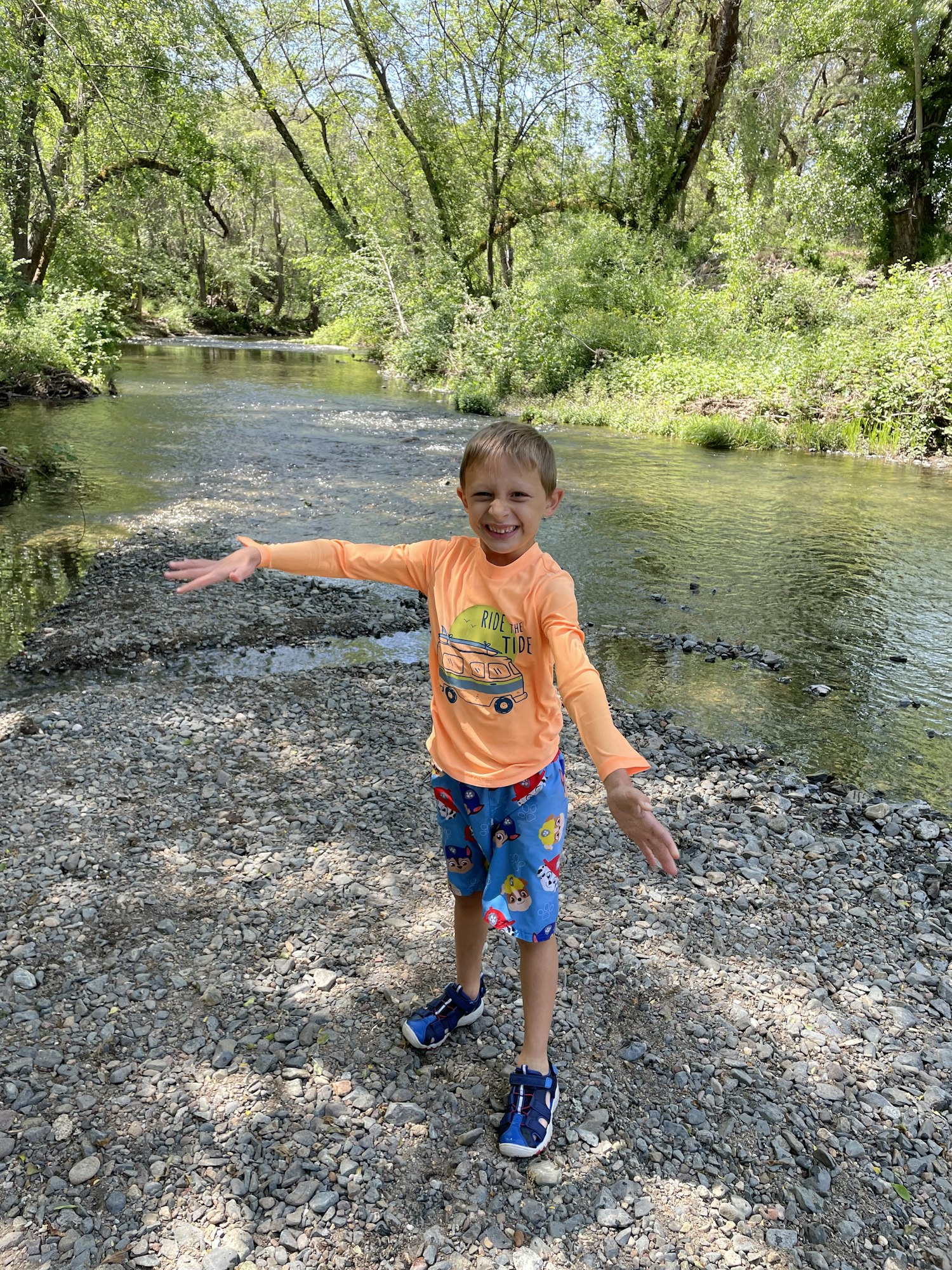 A child in colorful clothing stands smiling on a rocky riverbank surrounded by greenery.