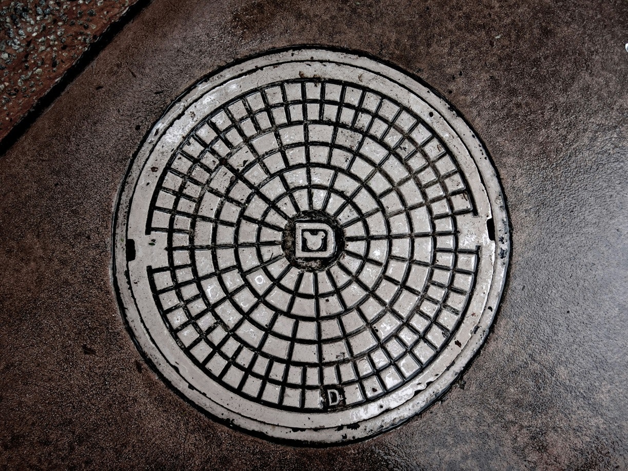 A weathered manhole cover with a geometric pattern on a concrete surface.