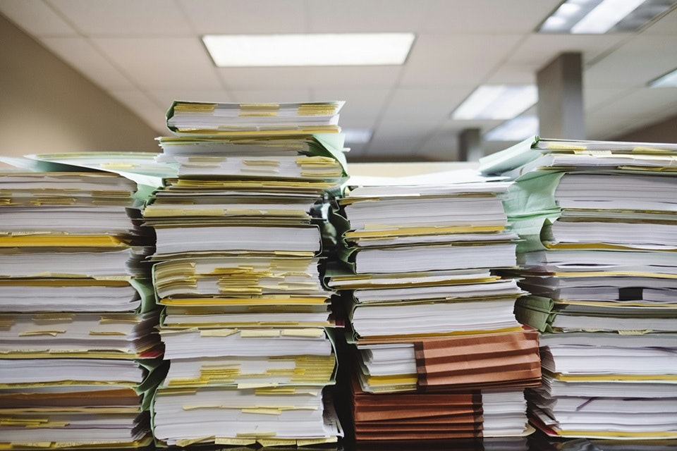 Large stacks of assorted papers and files on a desk, likely indicating a busy work environment.
