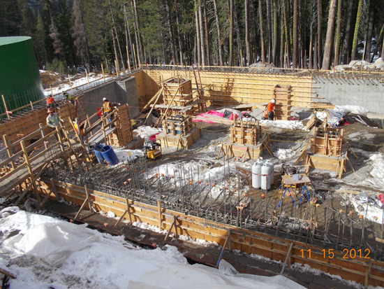 Construction site with workers, wooden frames, concrete, and equipment, surrounded by snowy ground and trees.