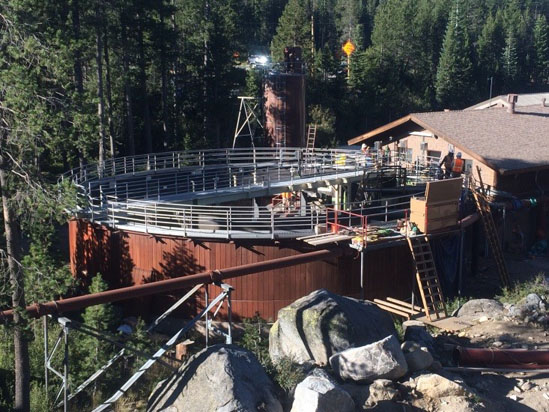 A large circular structure with railings, surrounded by trees and rocks, next to a building. Construction materials are visible.
