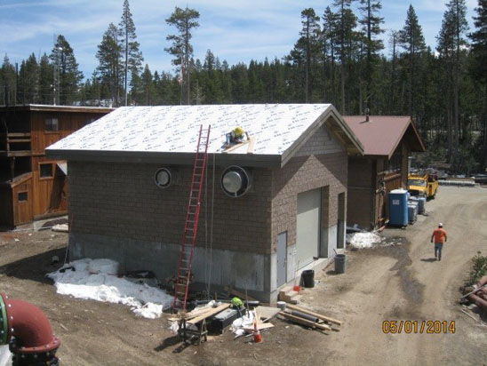 A small building under construction with scaffolding, a ladder, and materials around it. A worker is on the roof; trees in the background.