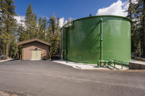 A green water storage tank next to a small building surrounded by trees under a clear sky.