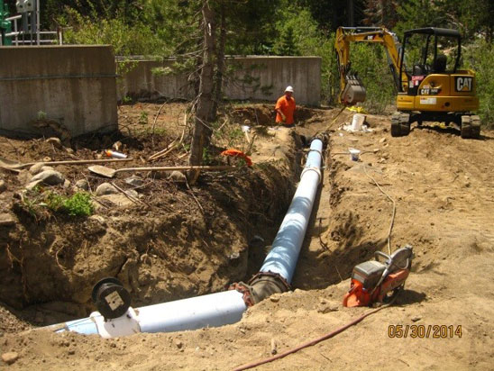 Construction site with a worker, an excavator, and large pipes being positioned in a trench.