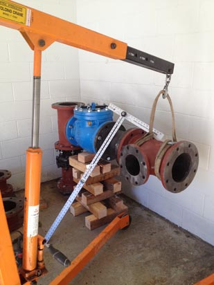 A hoist holds a red pipe fitting, with valves and stacked wooden blocks nearby.