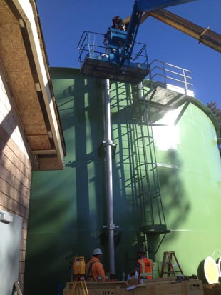 Workers with safety gear near a large green tank using a lift for maintenance or construction tasks.