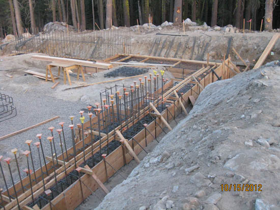 Construction site with rebar, wooden forms, and gravel foundation. Forest in the background.