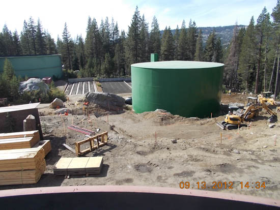 Construction site with a large green tank, wooden beams, and machinery, surrounded by trees.