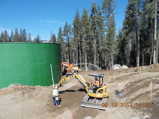 Construction site with a green tank, a worker, and a small excavator near a forest.