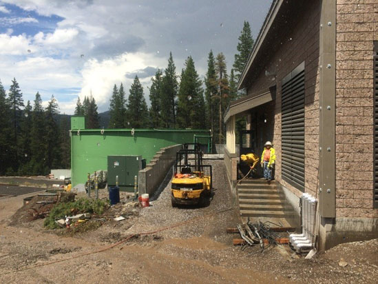 Construction site with workers, a forklift, and a large green tank surrounded by trees.