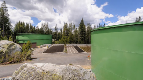 Water treatment facility with green tanks and basins, surrounded by forest under a partly cloudy sky.