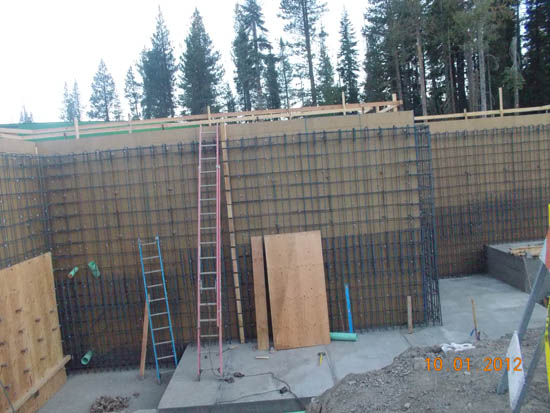 A construction site with wooden formwork, ladders, concrete foundation, and a forest in the background.