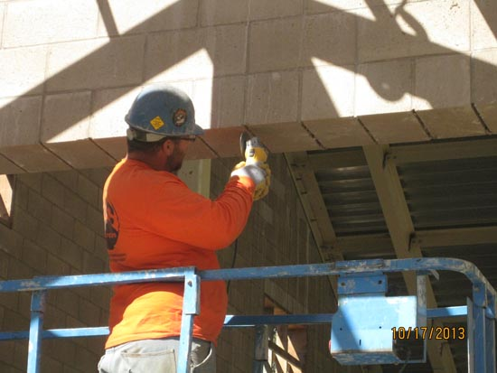 A construction worker in an orange shirt uses a power tool while standing on a lift.