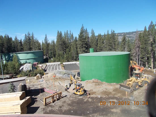 Construction site with two large green tanks, excavator, and workers surrounded by trees.