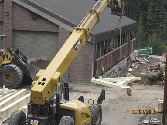 Construction site with a crane lifting a metal beam, workers nearby, and a building in the background.