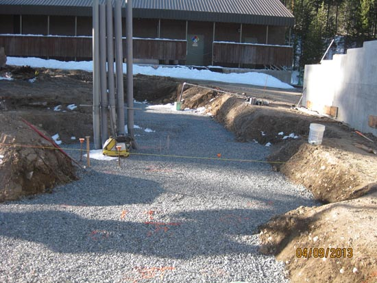 Construction site with gravel, trenches, pipes, building in the background, and snow near the perimeter.