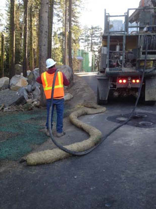 A construction worker in a safety vest and helmet operates a hose from a truck, possibly for landscaping or erosion control.