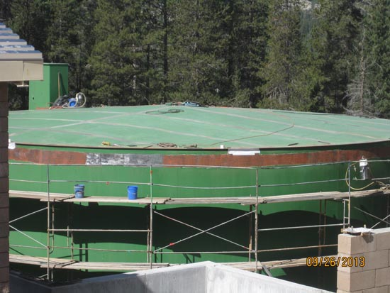 A large green water tank with scaffolding around it, amidst trees and equipment on top.