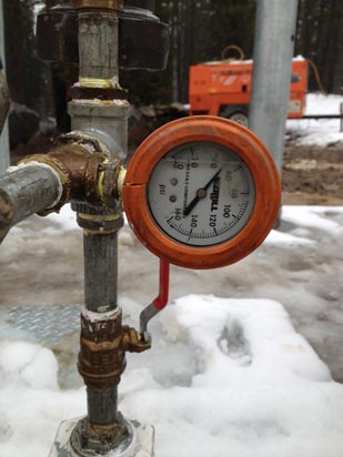 A pressure gauge attached to a pipe with a snowy background and construction equipment in the distance.
