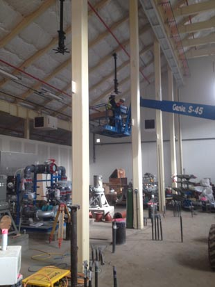 A construction site with equipment, pipes, and a worker on a Genie lift inside a large industrial building.