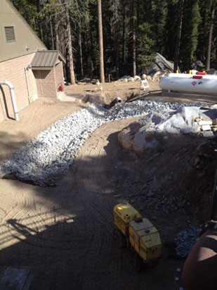 Construction site with a gravel path, yellow machinery, and a propane tank surrounded by trees.