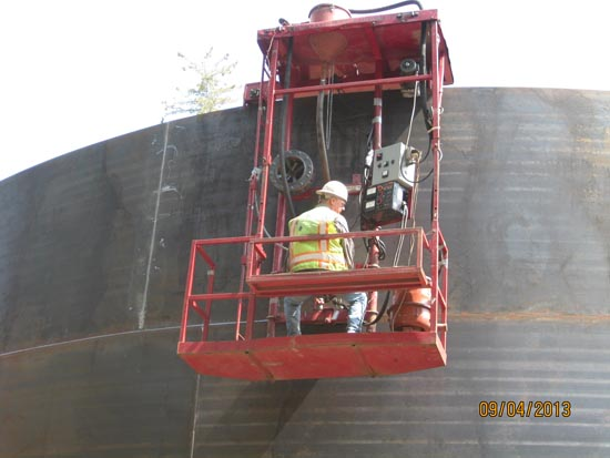 A worker in a hard hat and safety vest operates equipment inside a red lift basket near a large metal structure.