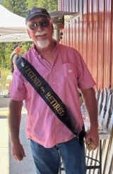 A man wearing sunglasses and a "The Legend Has Retired" sash, standing outdoors in casual attire.