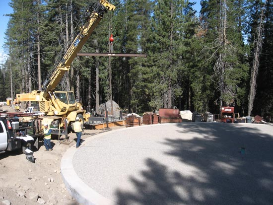 A crane lifting a beam at a construction site in a forest area with workers and vehicles nearby.