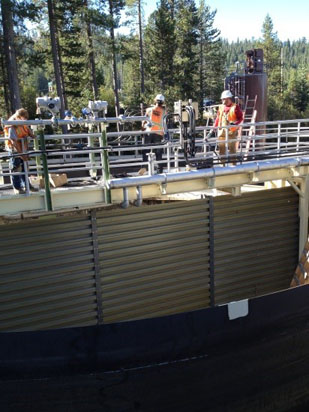 Workers in safety gear stand on a large industrial structure surrounded by trees.