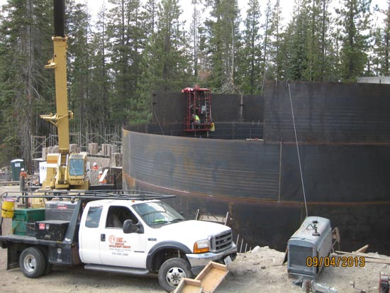 Construction site with a crane truck, large metal structure, and workers in a forested area.