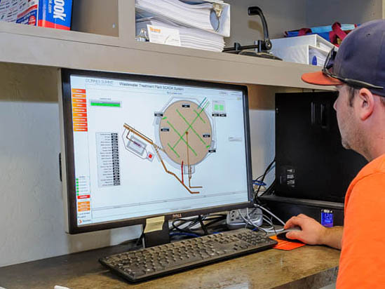 A person in an orange shirt uses a computer displaying a wastewater treatment plant SCADA system interface.