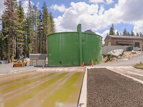 A large green storage tank on a construction site with surrounding trees and buildings.