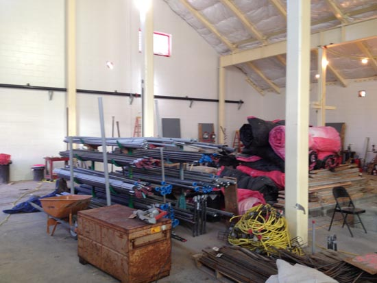 A construction site with stacked pipes, pink insulation rolls, cables, a wooden crate, and scattered tools in a large building interior.