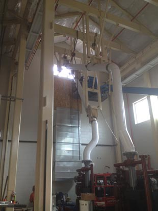 Industrial setting with large machinery, pipes, and overhead structures inside a tall building.