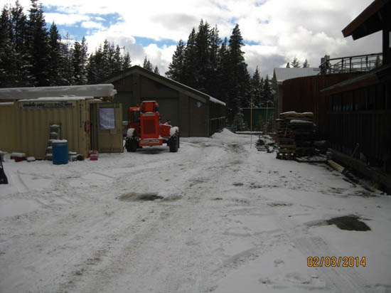 A snowy outdoor area with a red vehicle, containers, wooden structures, and trees in the background.