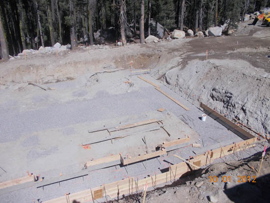 Construction site with a gravel foundation, wooden frames, and surrounding forest.