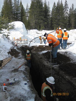 Construction workers in safety gear digging a trench in a snowy area near a forest.