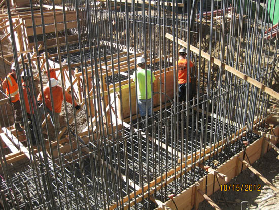 Construction site with workers wearing safety gear, surrounded by rebar and wooden framing.