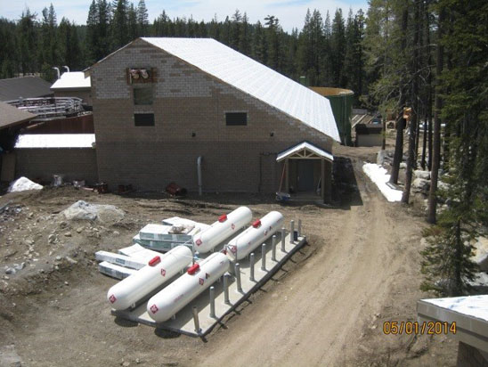 A construction site with a concrete building, propane tanks, and a dirt road surrounded by trees.