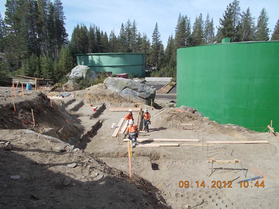 Construction site with workers, green storage tanks, and forest background.