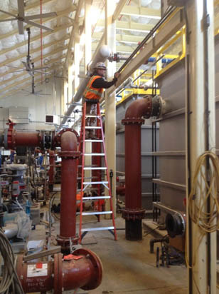A person in construction gear works on pipes from a ladder inside an industrial facility.