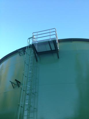 A large green industrial tank with a metal ladder and platform on its side, against a clear blue sky.