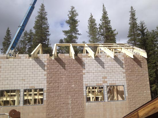 Construction site with a brick building, a crane, and roof trusses being installed, surrounded by trees.
