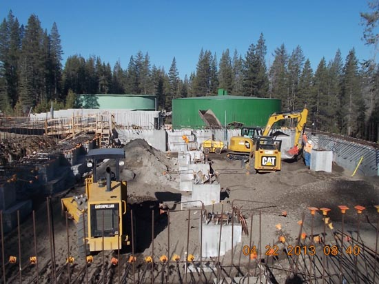 Construction site with machinery, concrete structures, and green tanks in a forested area.